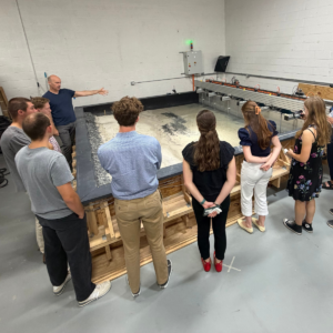 A group of national security policy center interns stand in front of a large, table-like wooden structure filled with grains of sand as a UVA researcher explains its significance
